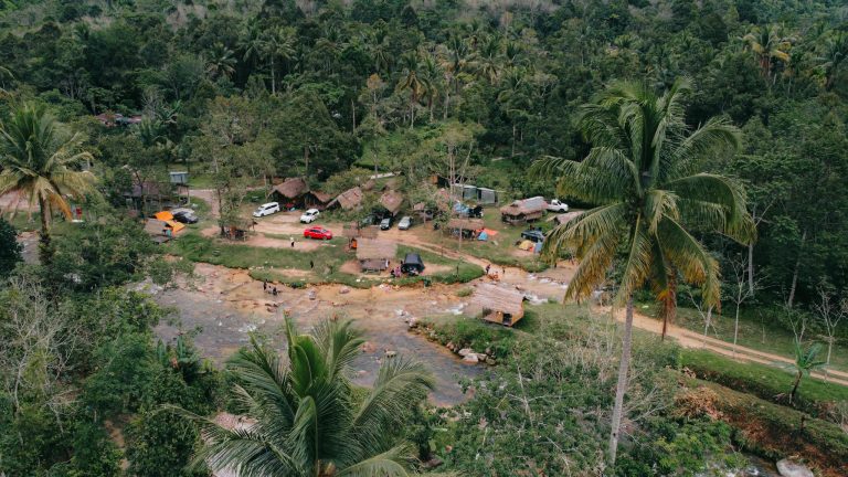 Aerial view of a tropical campsite by the river, showcasing tents, huts, and palm trees.